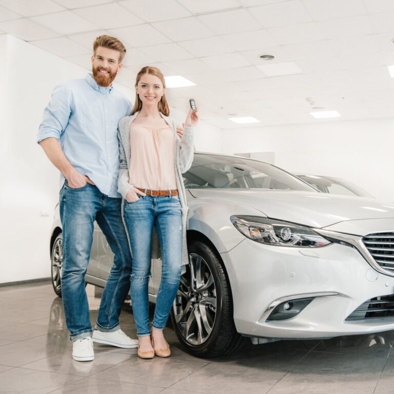 happy-couple-with-car-key-standing-at-car-in-dealership-salon-e1616384906835